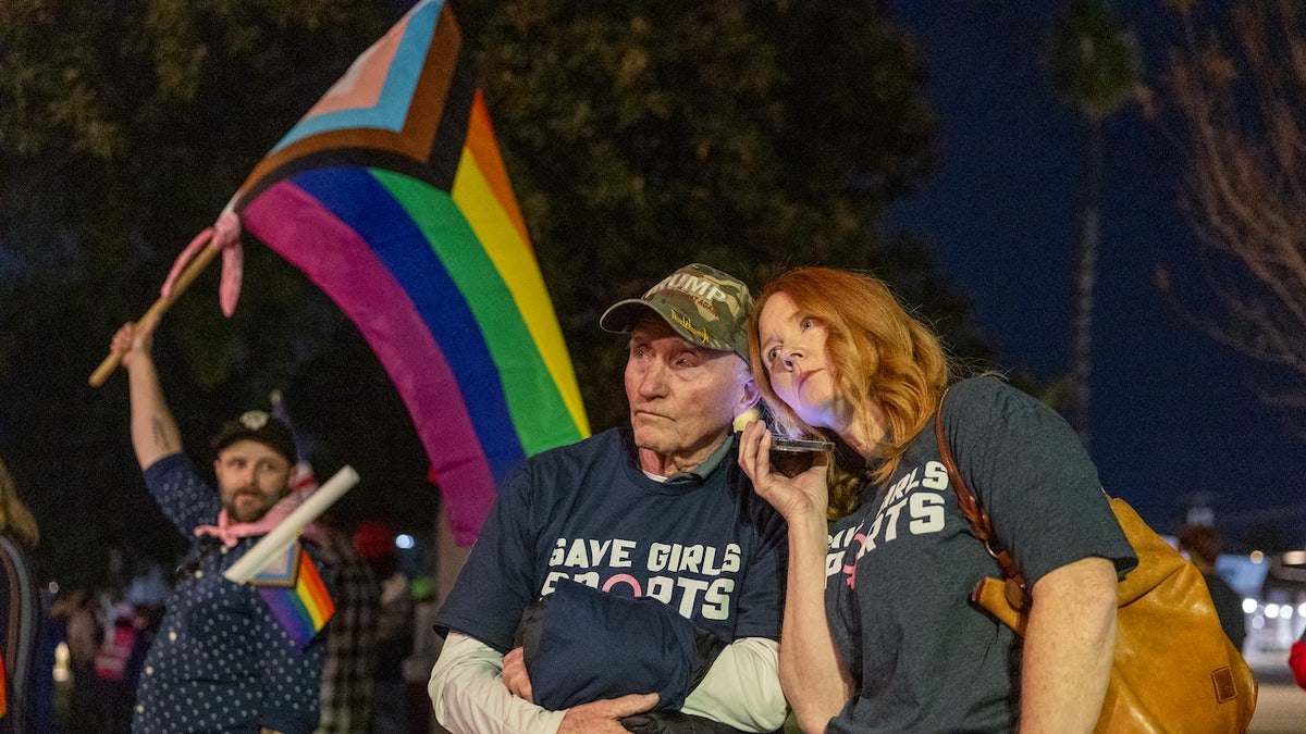 Transgender athlete supporter Kyle Harp, left, of Riverside holds the progress  pride flag as "Save Girls Sports" supporters Lori Lopez and her dad Pete Pickering, both of Riverside, listen to the debate as they join the overflow crowd converging outside the Riverside Unified School District meeting Thursday night to debate the rights of transgender athletes to compete in high school sports Thursday, Dec. 19, 2024. 