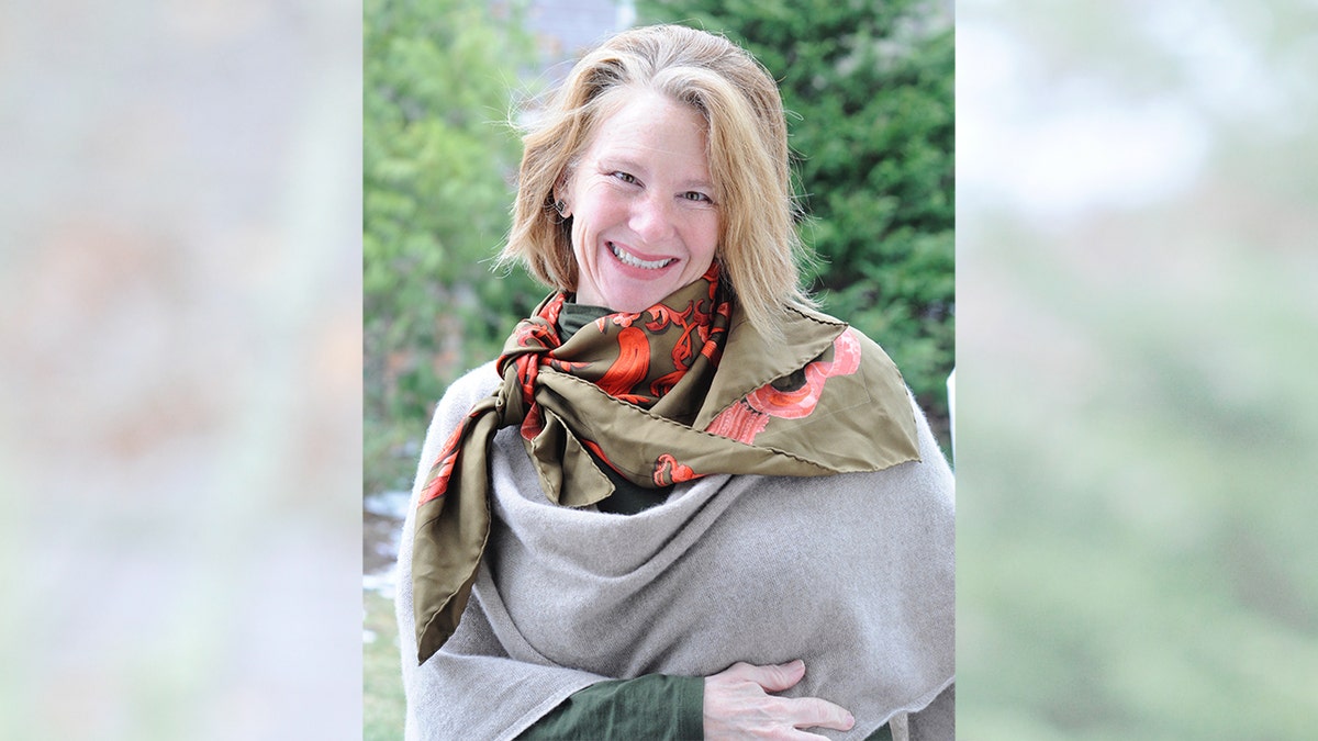 Lisa Brown Langley smiles while wearing a scarf and standing in a wooded area.