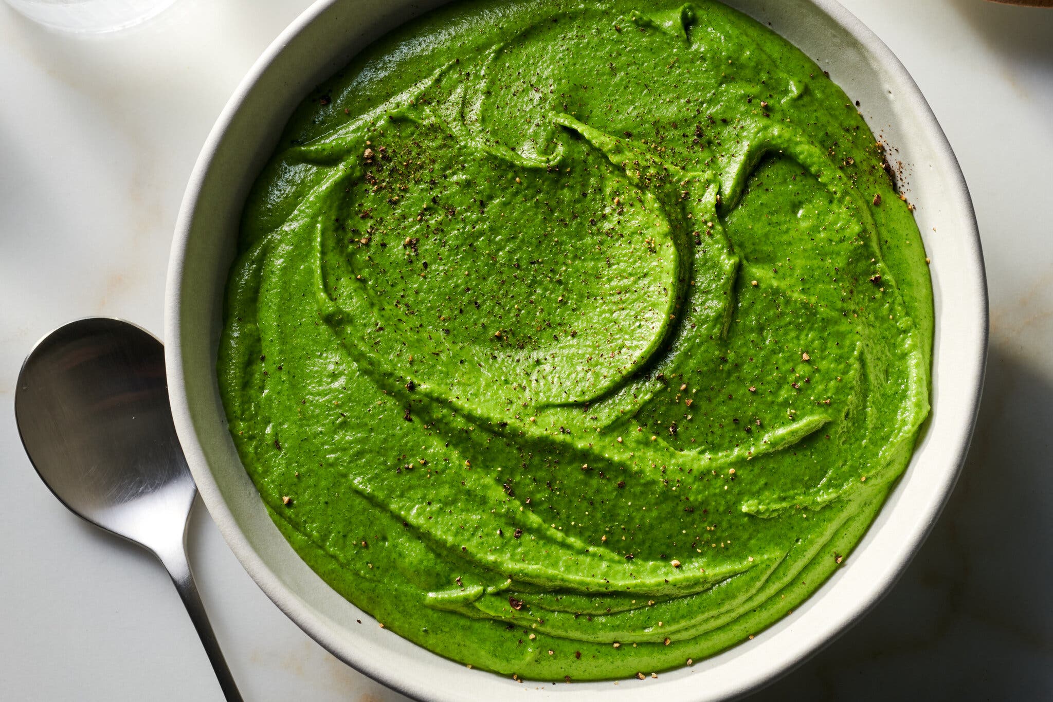 An overhead image of a white bowl filled with a bright green broccoli purée sprinkled with black pepper. To the left of the bowl there is a silver spoon.
