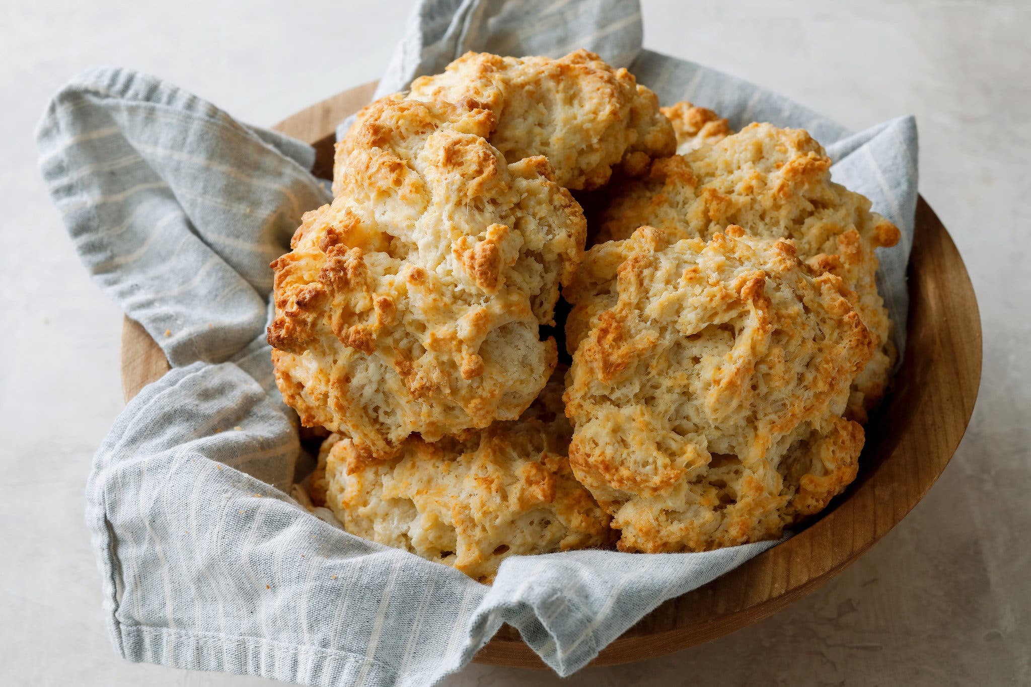 A side image of a wooden bowl lined with a light blue and white striped dish towel filled with golden brown, craggy drop biscuits.