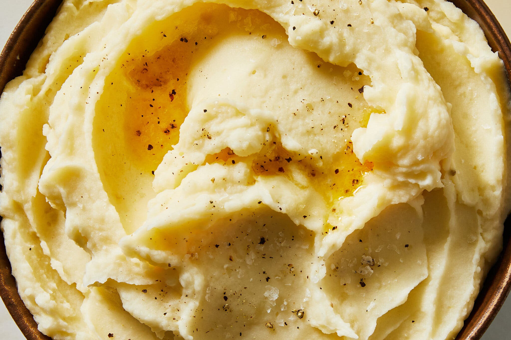A close-up image of mashed potatoes dotted with black pepper and topped with melted butter in a brown bowl.