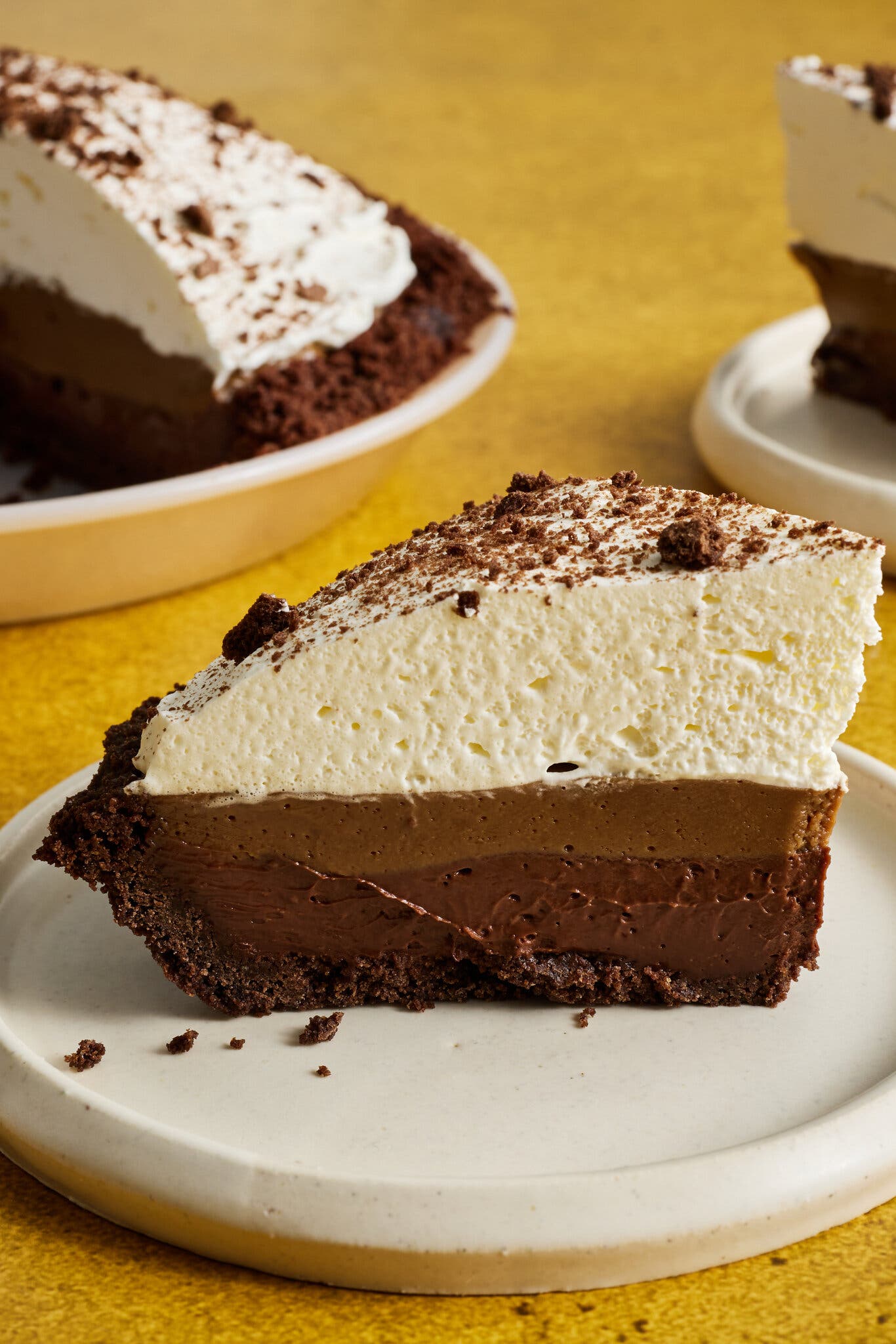 A slice of creamy chocolate and espresso pudding pie with a thick whipped-cream topping sits on a white plate, with the rest of the pie sitting half in frame behind it.
