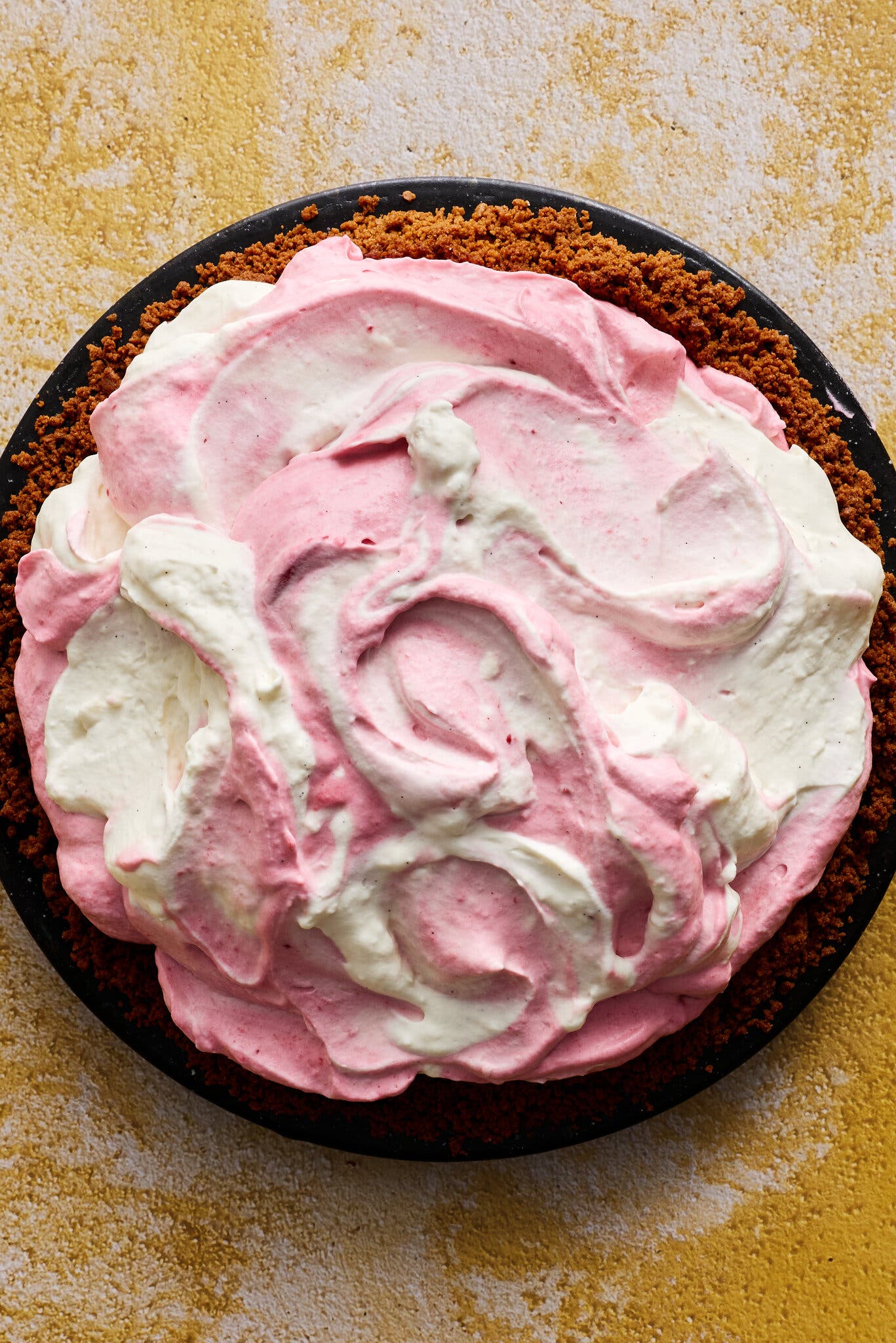 An overhead shot of a pie with a crumbly brown crust and dolloped-in swirled pink and white mouse sitting on a yellow and white background.
