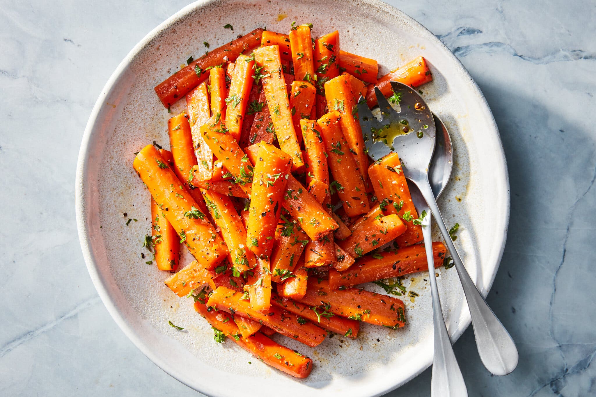 A light plate is full of bright orange carrots, coated in oil and green herbs. A silver serving spoon and fork rest in the dish.