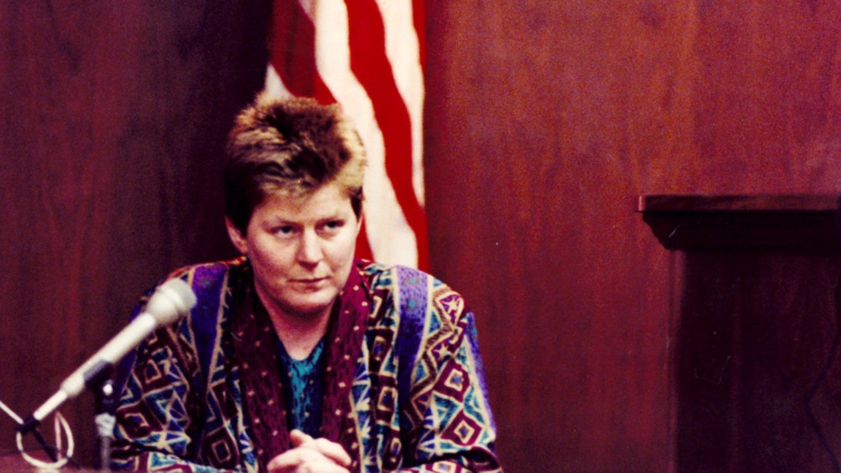 Tyria Moore wearing a multicolored blouse, sitting in front of an American flag in a courthouse.