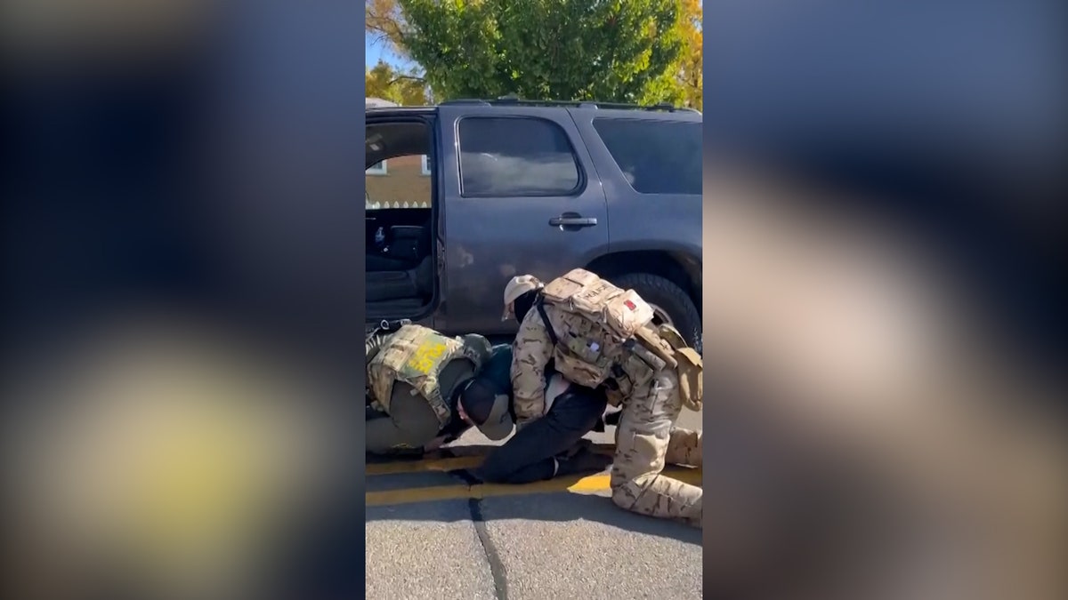 Two agents pin down a suspect in front of a patrol vehicle.