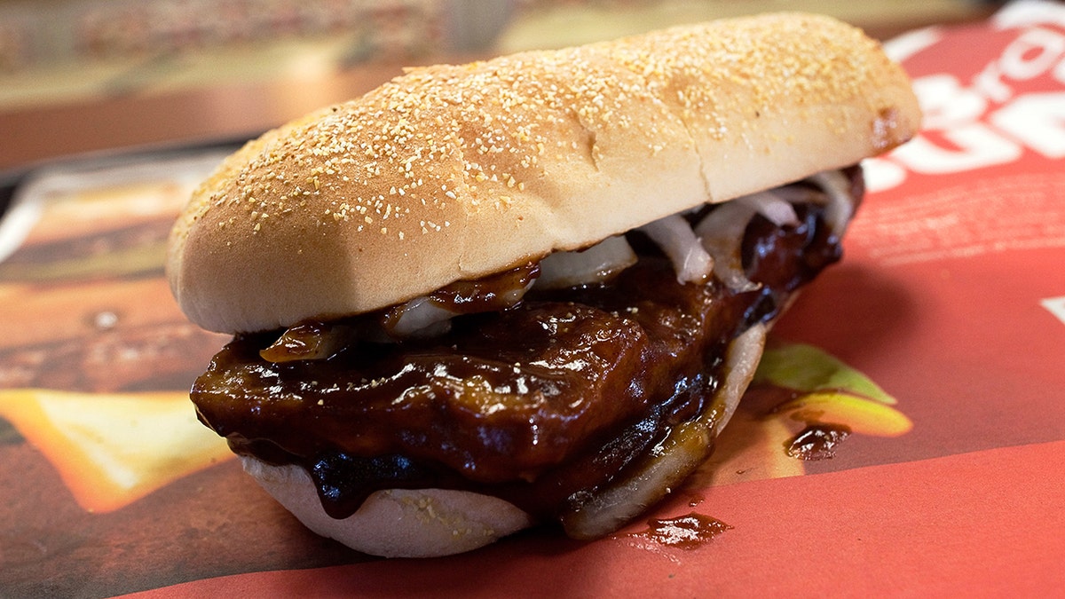 McRib sandwich seen up close on restaurant paper placemat; onions seen under break and dripping BBQ sauce.