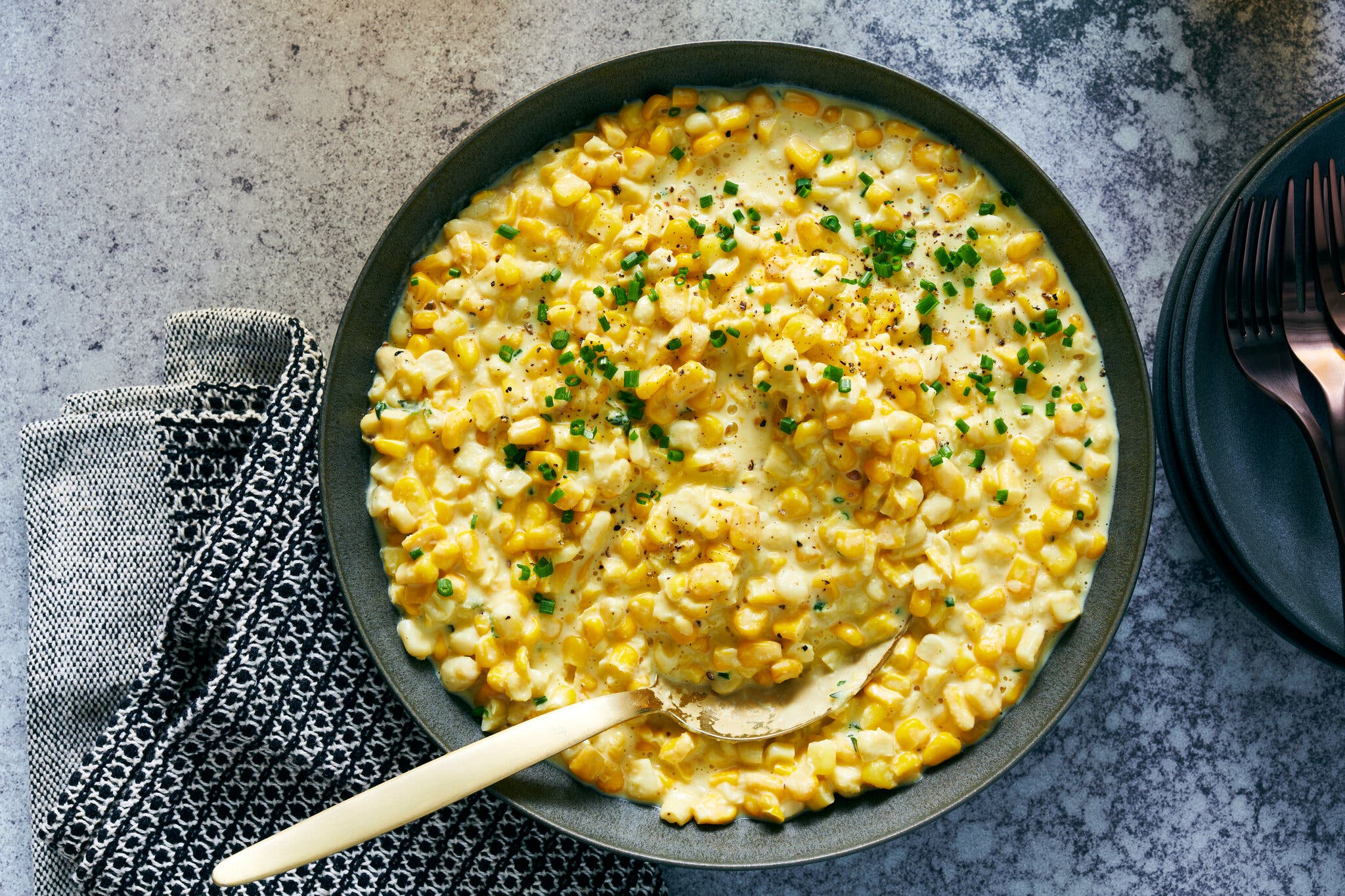An overhead image of a black serving bowl filled with creamed corn sprinkled with chopped green scallions. A gold serving spoon is tucked into the corn. A black and white dish towel is tucked under the plate. Peeking in to the right side of the frame is a stack of black plates with a stack of gold forks.