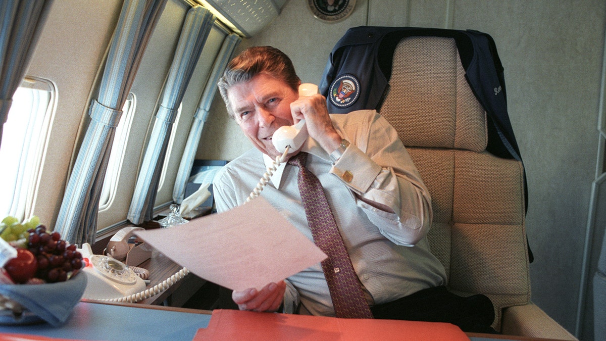 President Reagan making call from rotary phone, smiling, holding paperwork, aboard Air Force One.