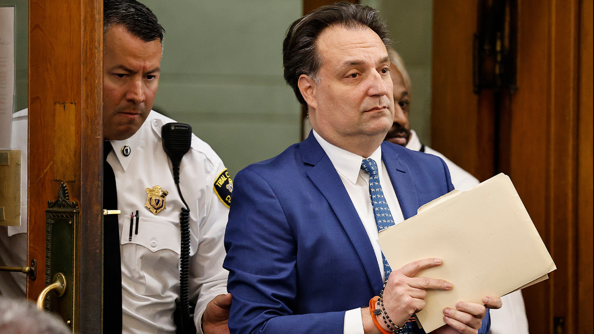 Brian Walshe enters the courtroom in a blue suit, with shackled wrists and carrying paperwork.
