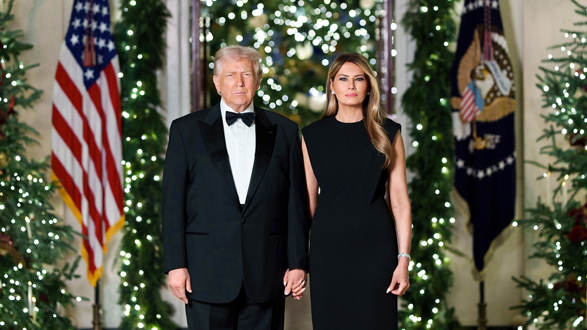 President Donald Trump and First Lady Melania Trump stand side-by-side in front of Christmas decor and two flags.