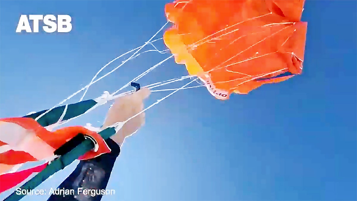 A skydiver grips parachute lines as an orange reserve canopy inflates against a clear blue sky.