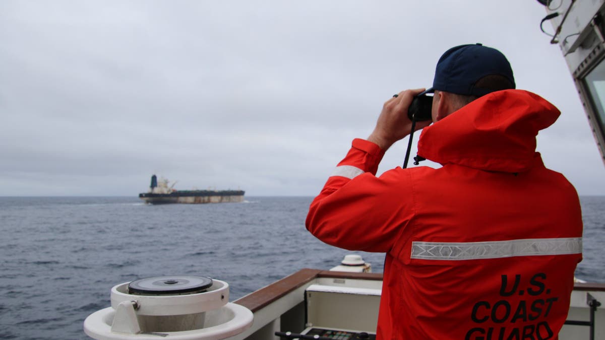 A member of the U.S. Coast Guard looking through binoculars at an oil tanker