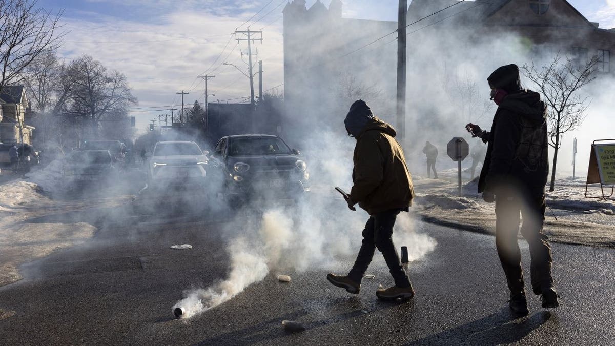 Tear gas canister smokes on a Minneapolis street as anti-ICE agitators walk nearby during an enforcement operation.