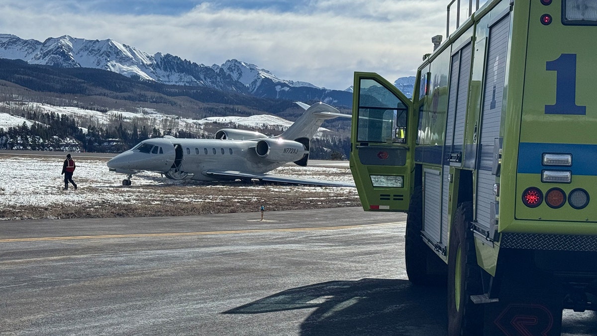 A jet rests off a runway on snow-dusted grass at a rural mountain airport as emergency crews respond.