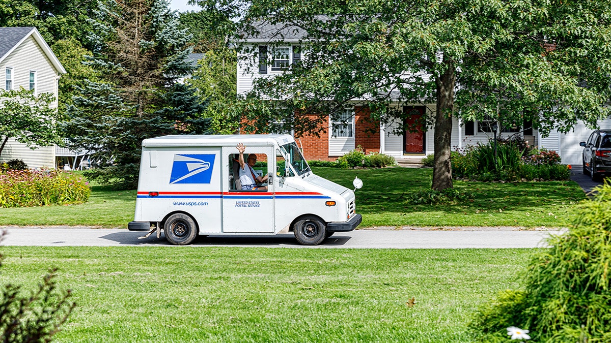 USPS mail carrier driving a postal delivery truck through a suburban neighborhood with trees and homes.