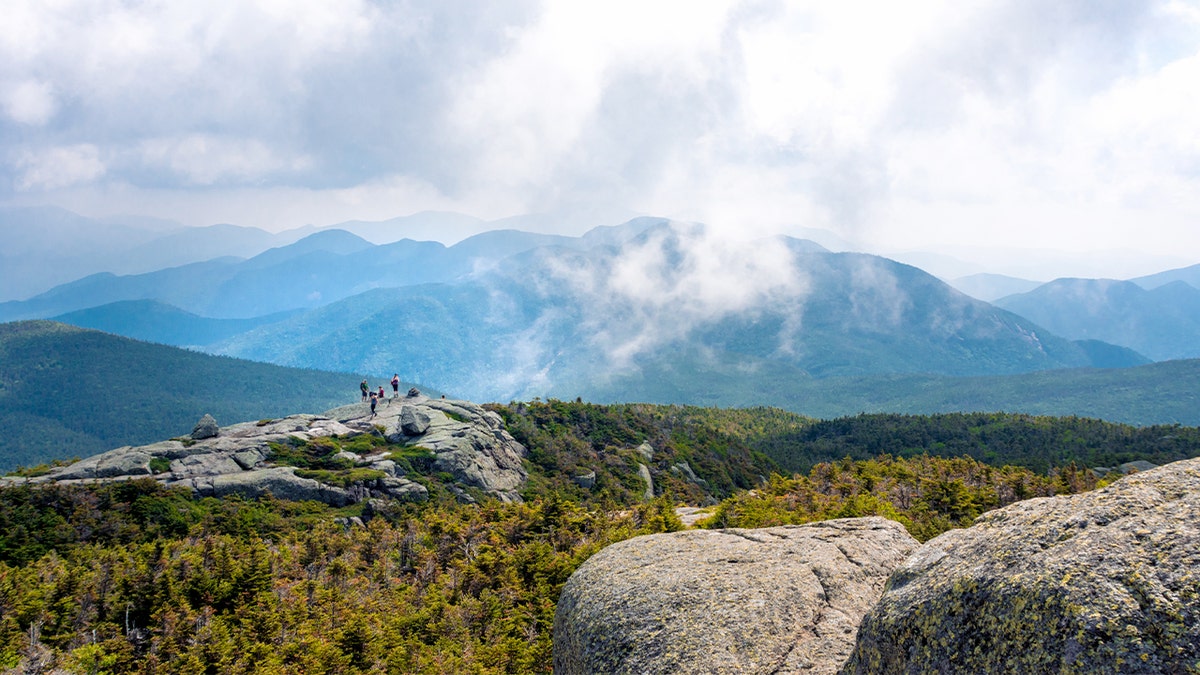 Hikers pause near a rocky summit in the Adirondacks, taking in sweeping mountain views under a clear winter sky.