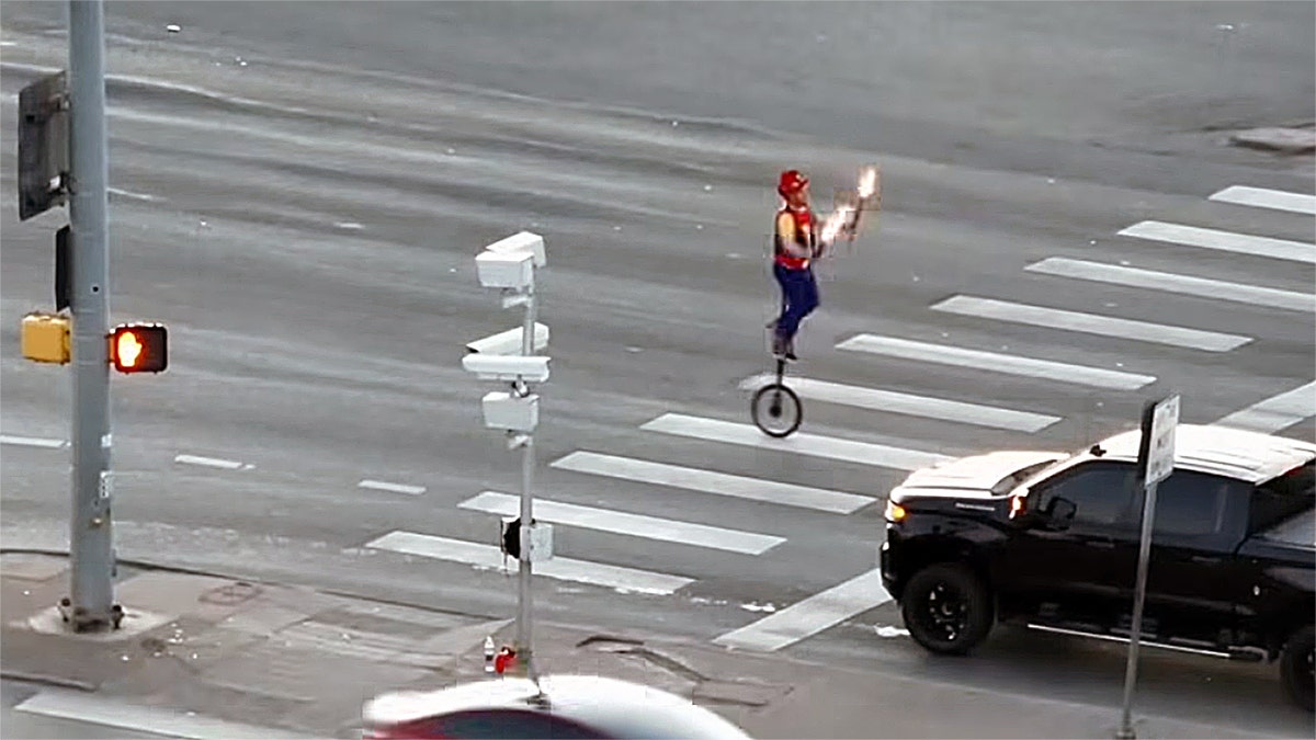 Fire-juggling unicyclist rides through a crosswalk at a busy intersection