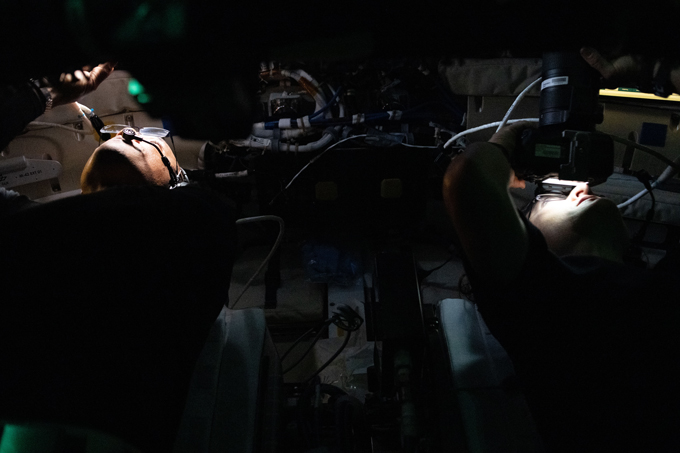 Two astronauts look up in a dark spacecraft. The only light is on their faces. Both wear glasses and hone is holding a camera.