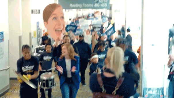 GIF of Mallory McMarrow walking into Democratic convention with marching band and giant face cut-out