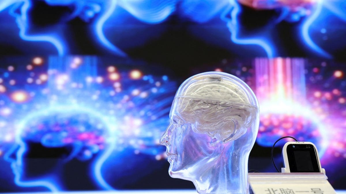 A translucent model skull displays a brain-computer interface in front of a colorful purple and blue background image of a human brain with neurons