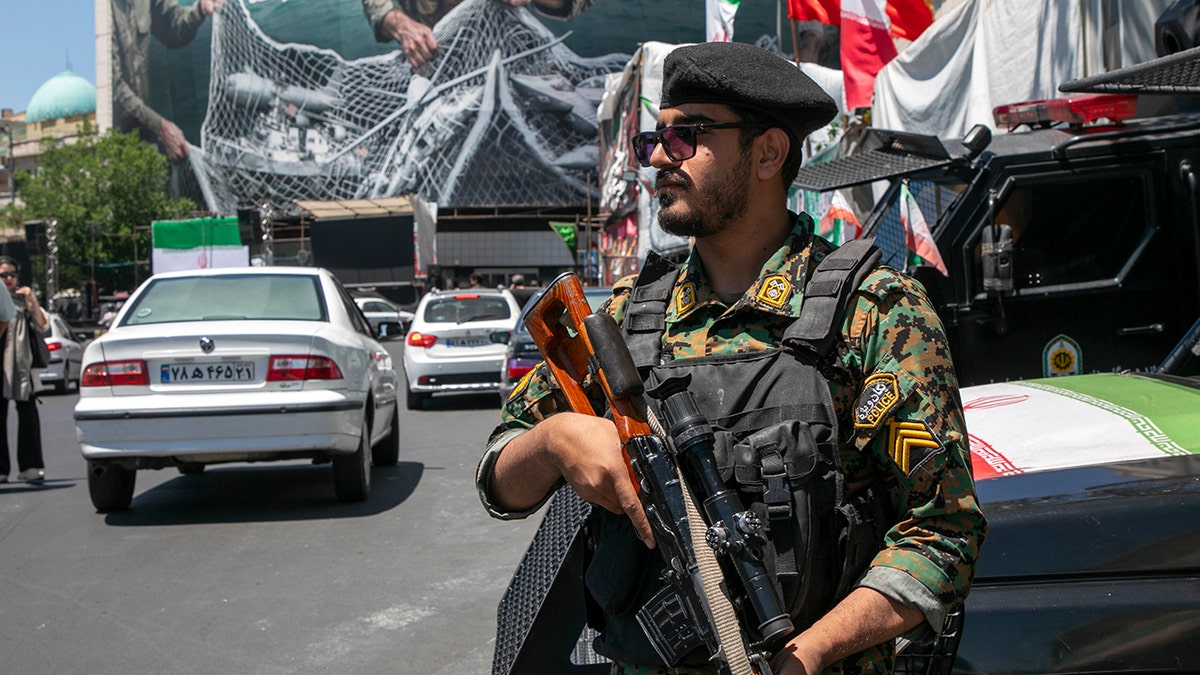 Iranian police officer standing on patrol near a poster depicting Iranian soldiers and U.S. military aircraft in Tehran