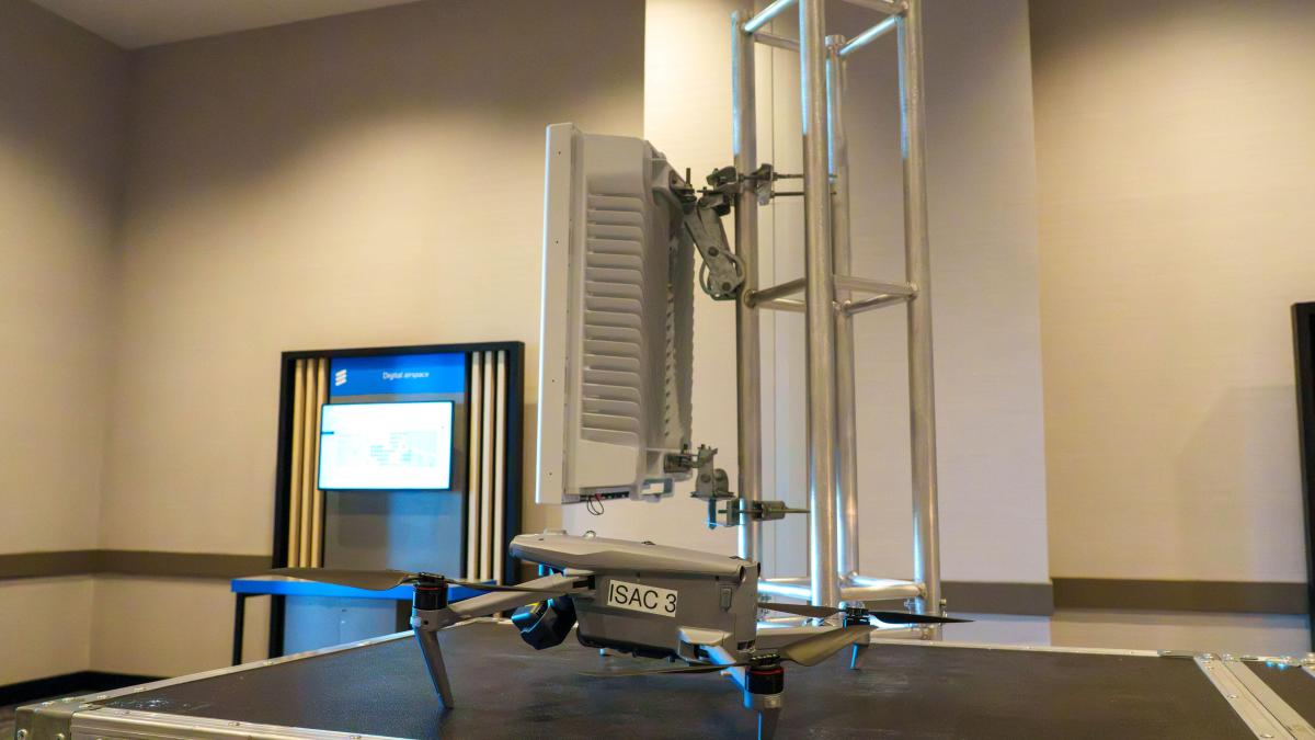 A drone sits on top of a case. In the background is a rectangular beige box mounted to a metal frame.