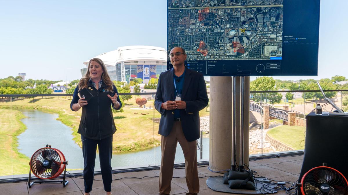 Two people speaking next to a large video monitor and a stadium in the distant background.