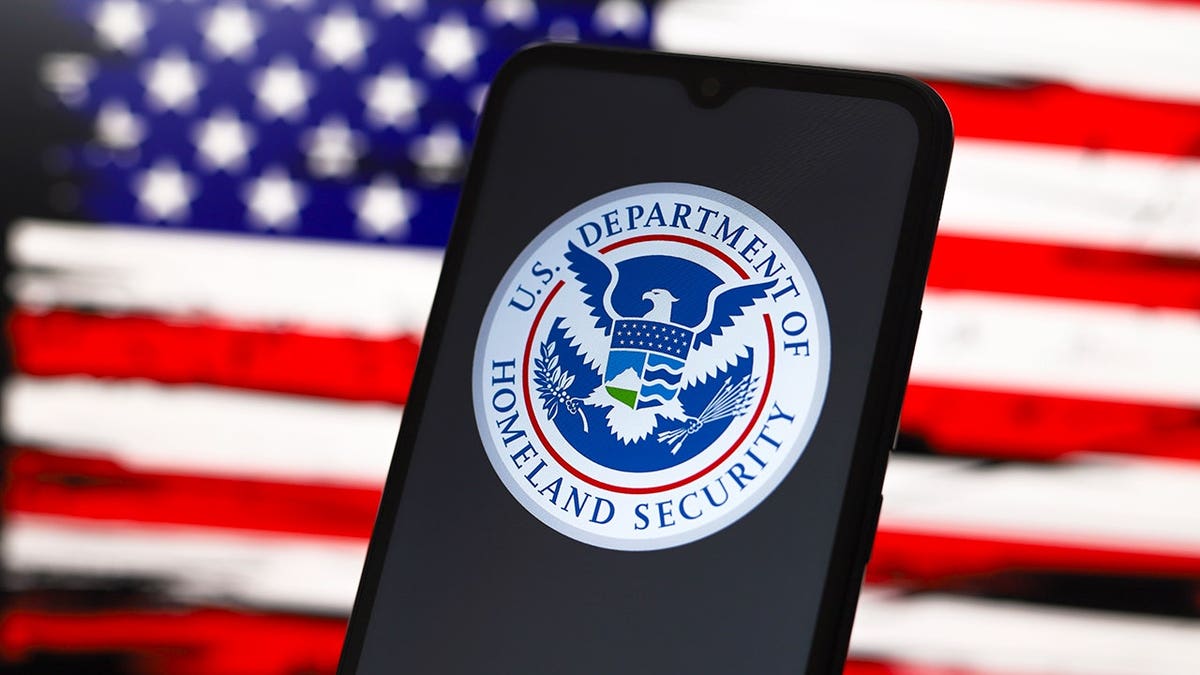 Department of Homeland Security logo on smartphone screen with American flag design on computer screen in background.