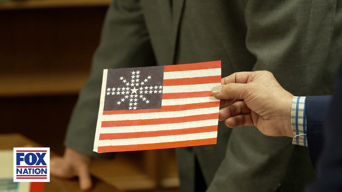 A hand holds a proposed 50-star American flag design with the stars arranged in a starburst pattern, one of the many submissions considered before the current U.S. flag was adopted.