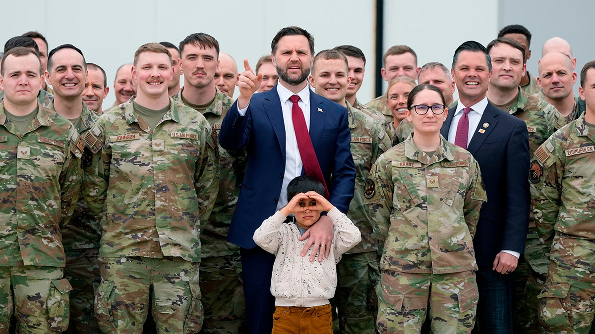 JD Vance points upward while standing with his son and military members in camouflage uniforms.