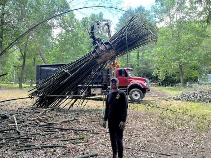 A man stands in front of a work trucj with a red cab. Above his head, a claw-like tool grasps a large bundled of recently cut bamboo stalks. Other stalks are scattered on the ground.