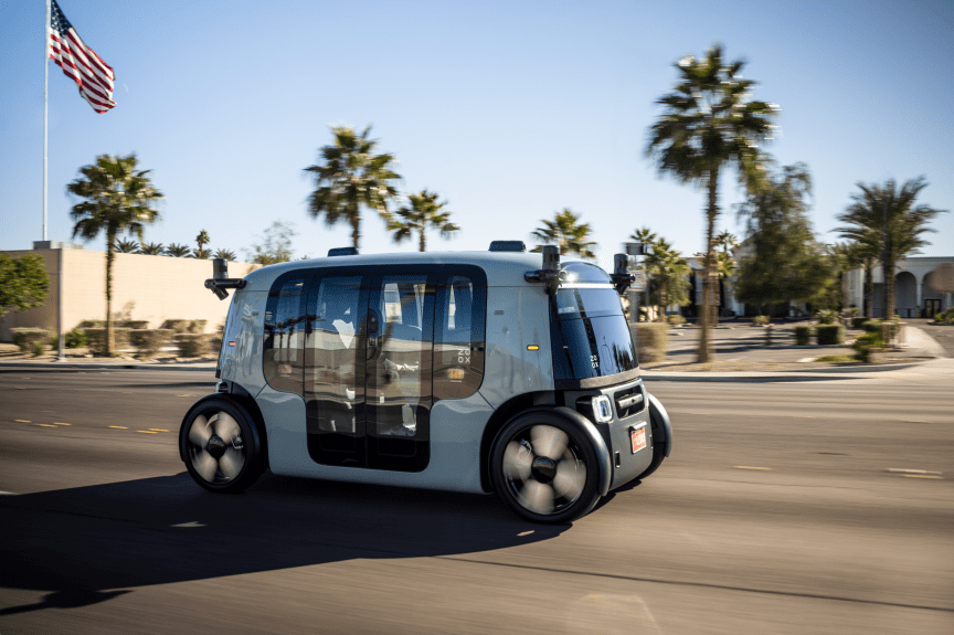 A Zoox robotaxi on the road in Las Vegas, with palm trees and an American flag in the background