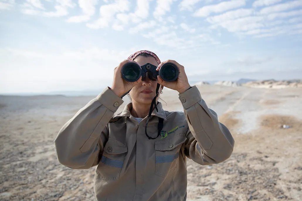 A frontal view of Cárdenas-Alayza looking through a pair of binoculars. The rocky coast behind her blurs as it stretches into the distance.