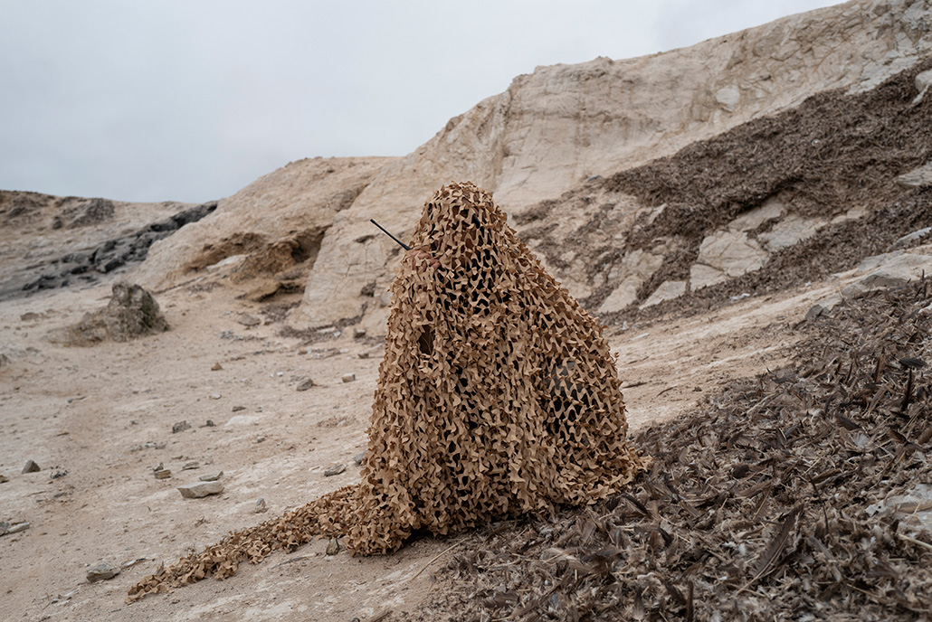 A human shape covered by camouflage netting on a coastline