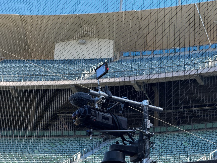 A phone mounted above serious cameras in front of netting, and far behind, fan seats.