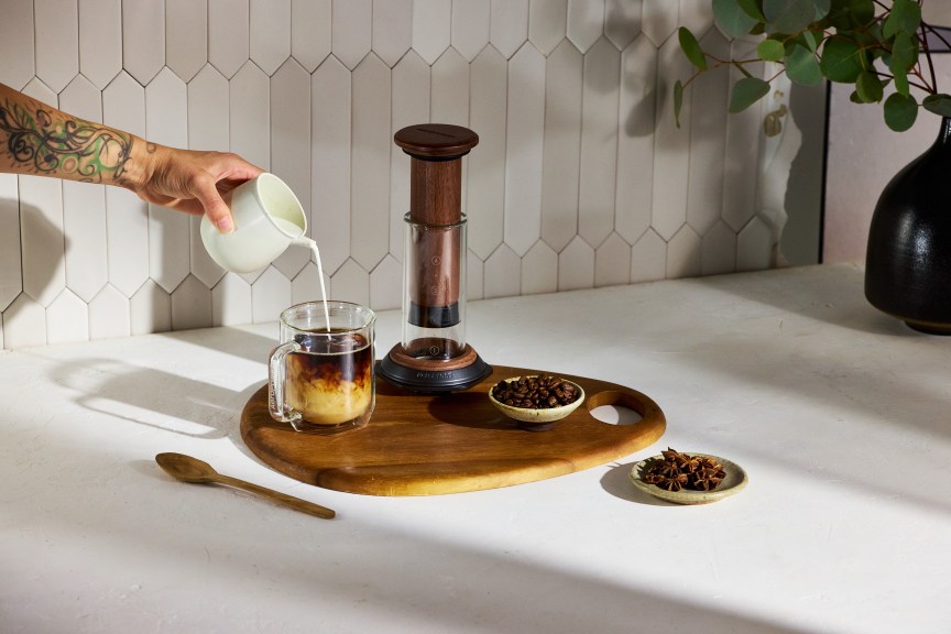 The walnut AeroPress on a walnut board next to a cup of coffee with milk being poured into it.