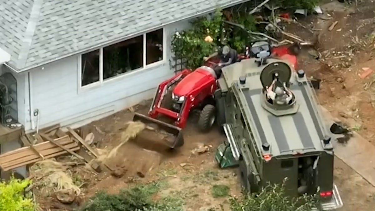 Armored SWAT vehicle rams a red tractor beside a residence.